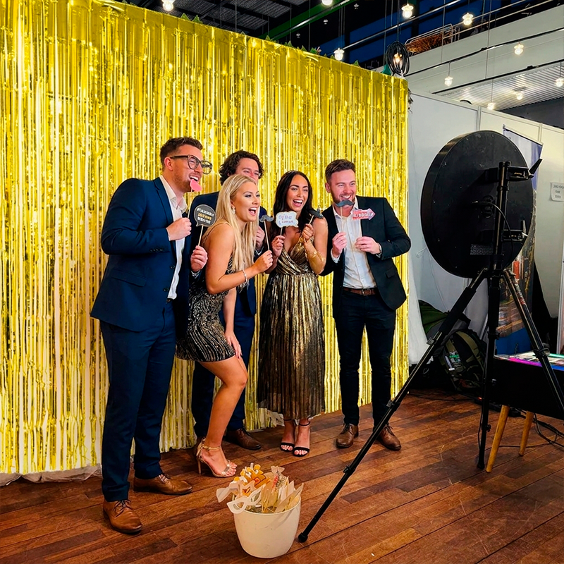 Fem gæster griner og poserer med rekvisitter foran en guldfarvat frynsegardin ved en SelfieCircle photo booth. Maskinen ses til højre på stativ med ringlys.