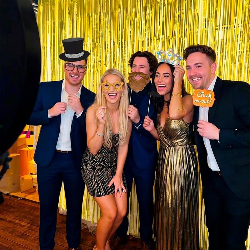 Fem gæster poserer og griner foran en gylden frynsegardin ved en photo booth. De holder rekvisitter op: hat, stjerne-briller, bart, krone og et skilt med "Check me out!". Festtøj i sort, guld og marineblå.